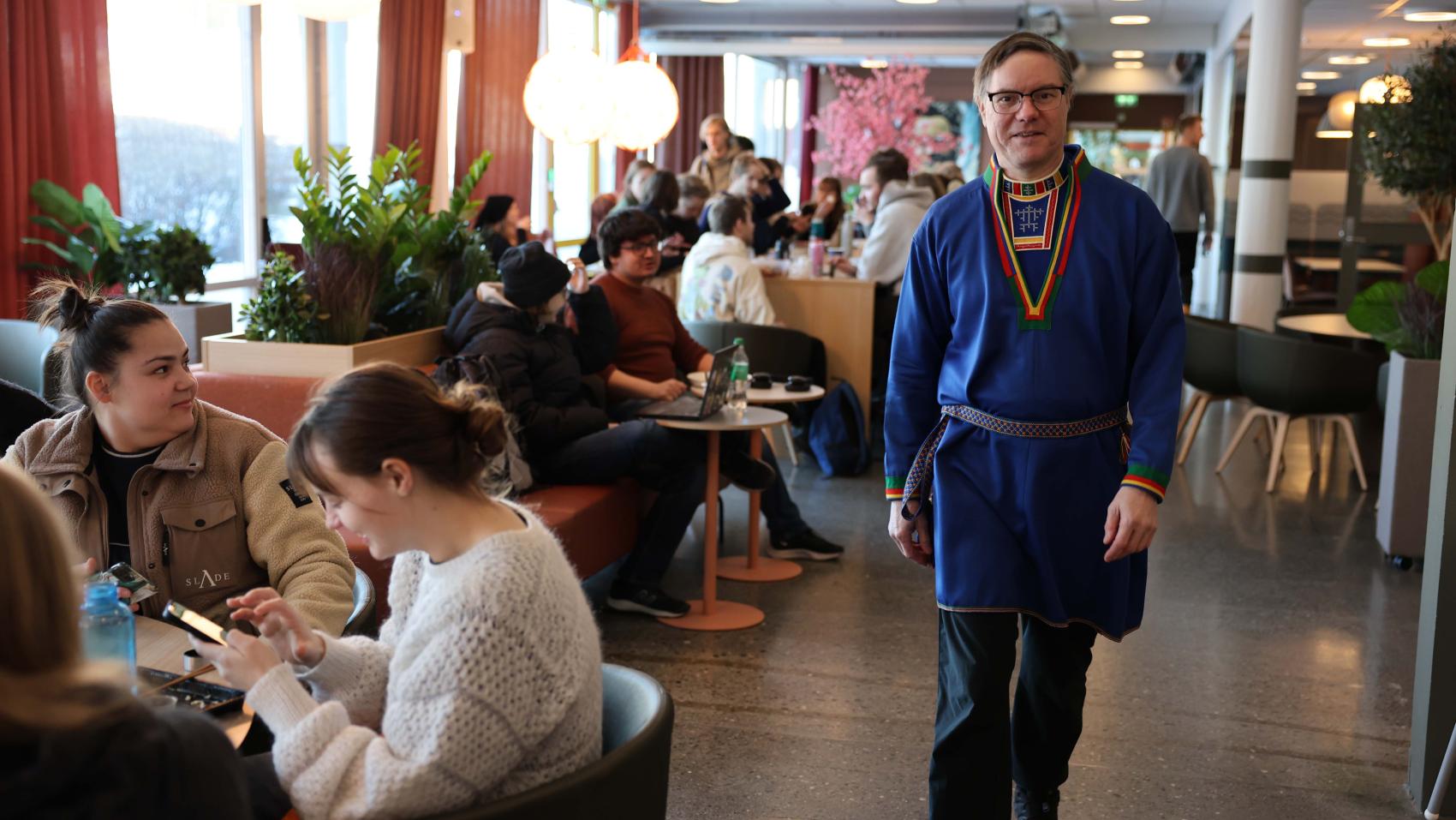 Mann i samisk kofte som går mot kamera inne i ei kantine hvor det sitter mange andre mennesker rundt bordene. Foto