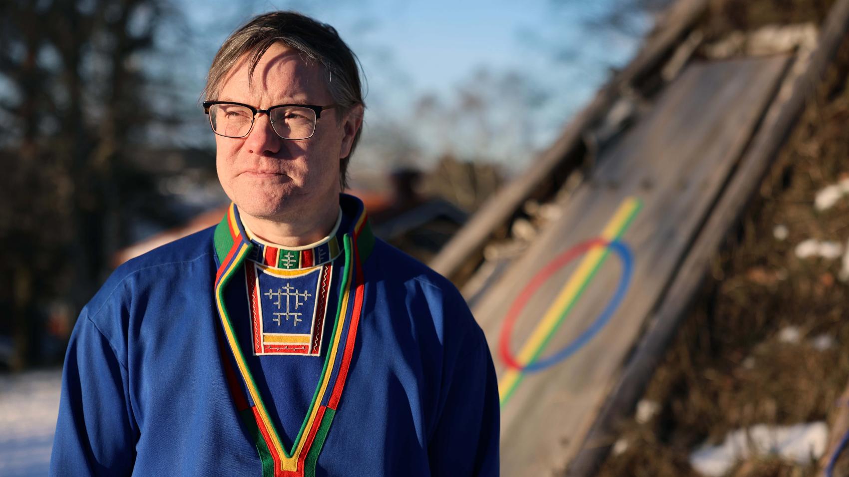 Nærbilde av mann i samisk kofte foran ei gamme. Foto