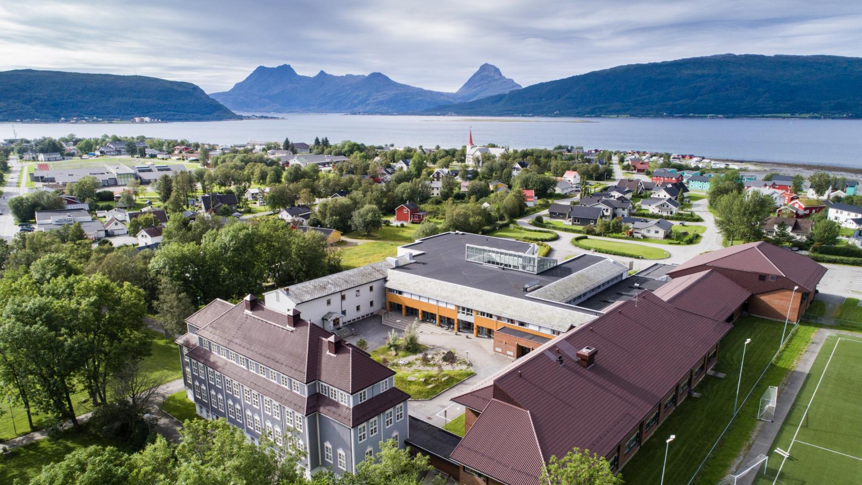 Landskapsbilde tatt med fugleperspektiv av flotte universitetsbygg i forgrunnnen, et tettsted i bakgrunnen, med fjorder og fjell i horisonten.