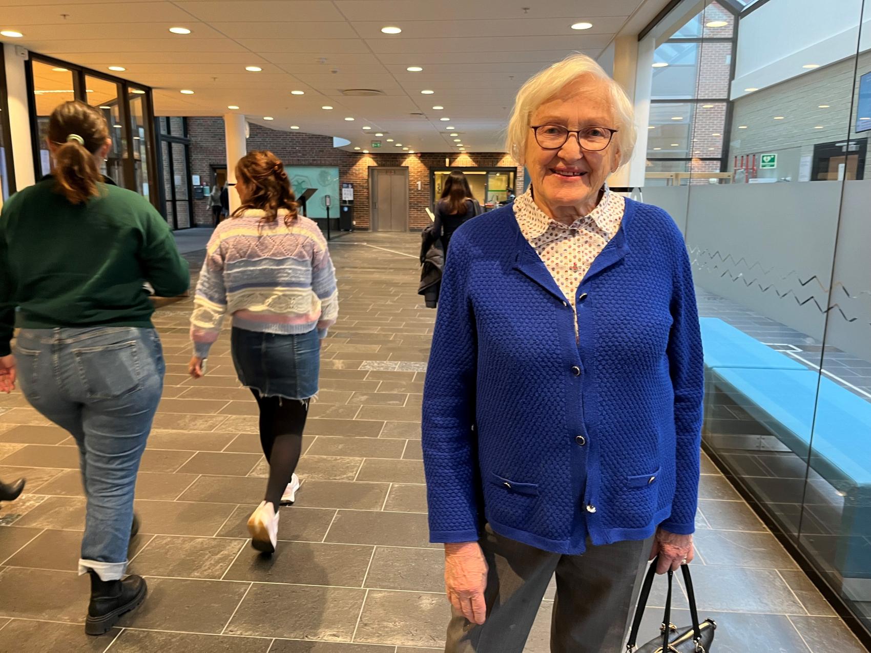 Eldre dame i korridor med gående mennesker bak. Foto