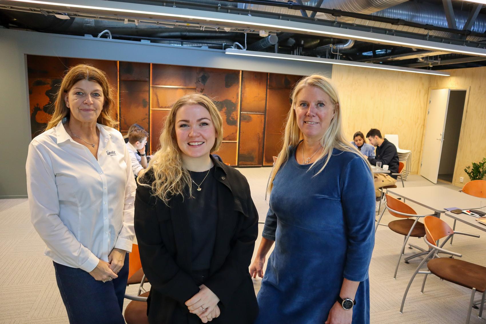 Tre smilende kvinner innendørs med fire personer som jobber i bakgrunn. Foto.