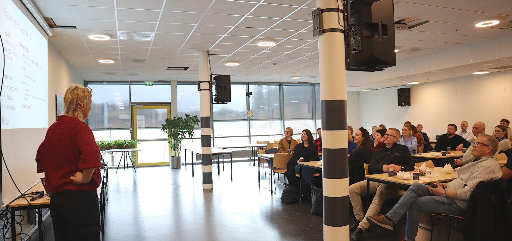 Kvinne kledd i rødt står foran skjerm, holder foredrag. Ca 20 personer sitter ved bord og ser på henne. 