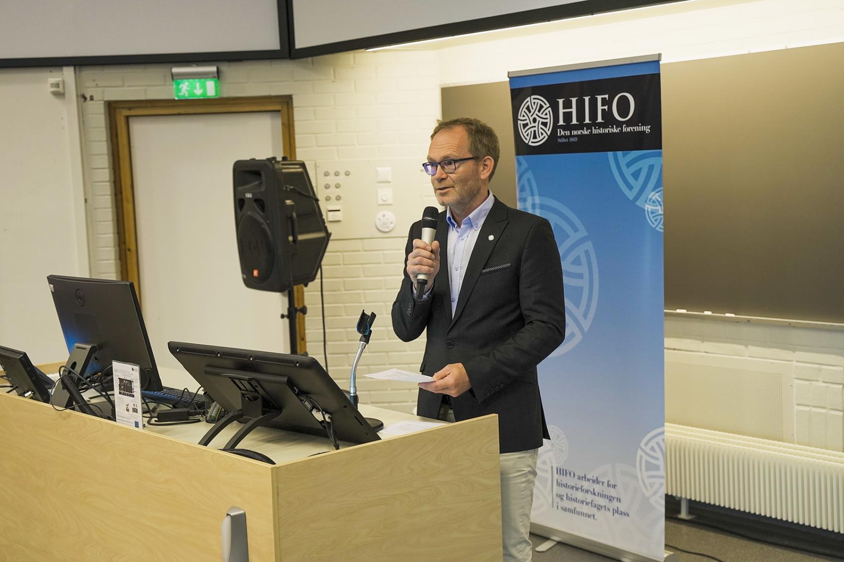 Mann i dress som snakkar i mikrofon. Foto