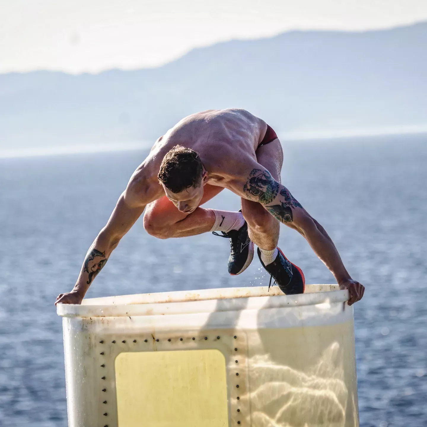 Mann i badebukse som hopper ned i et kar fylt av isvann med havet i bakgrunn. Foto