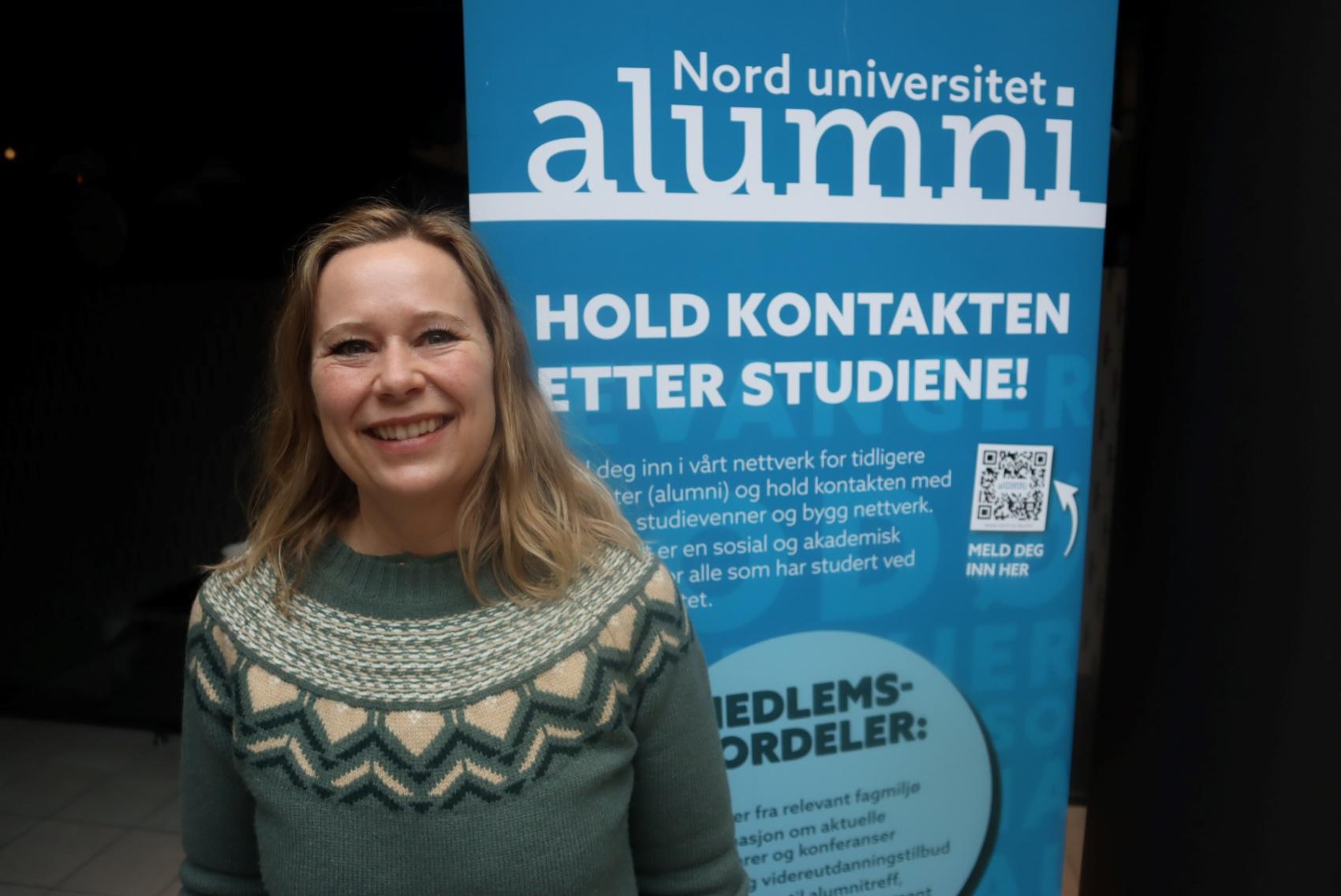 Smilende dame i mønstret strikket genser foran blå roll-up med alumni-info
