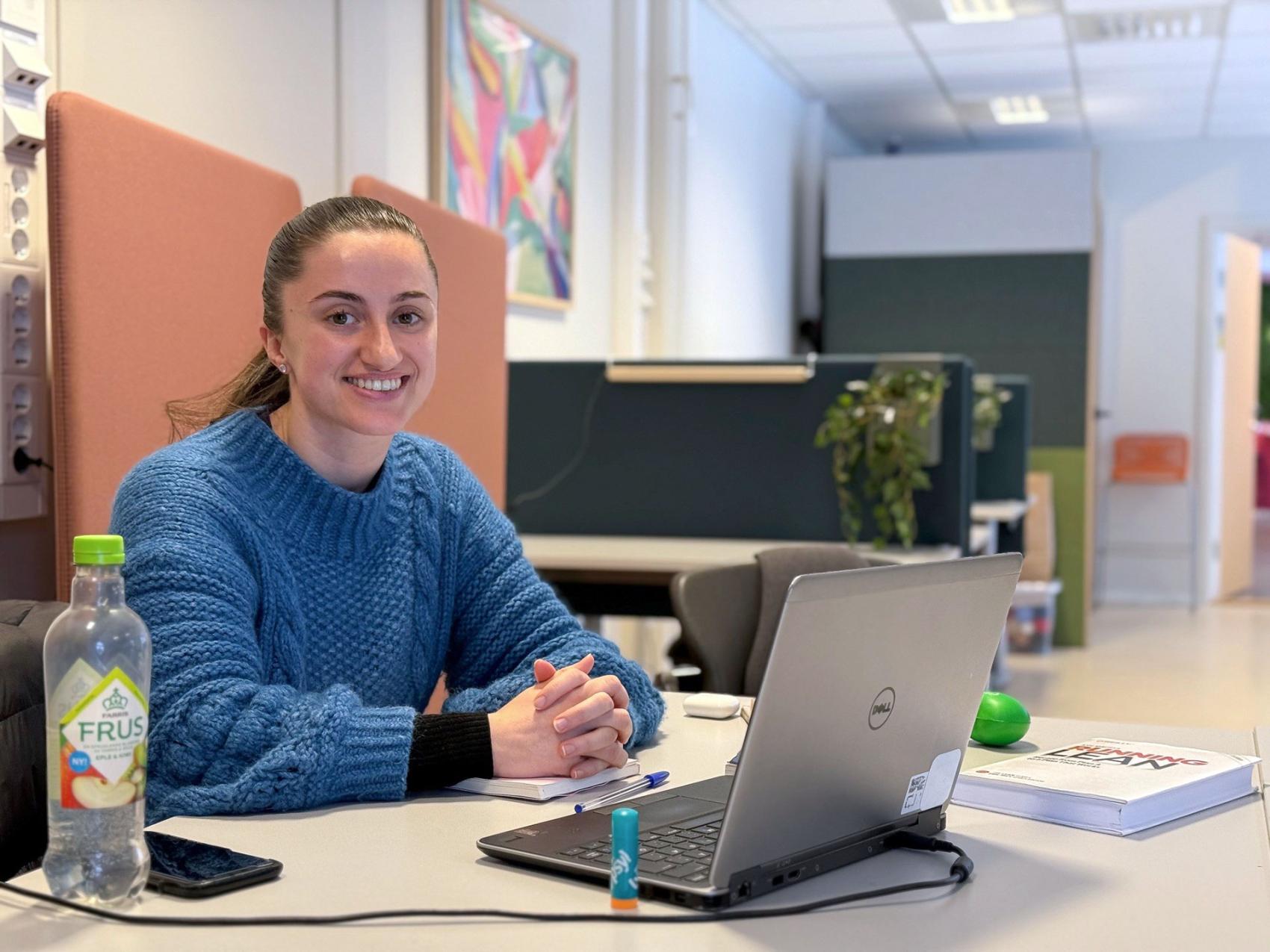 Ung kvinne med blå genser som sitter ved et bord med PCen oppslått.