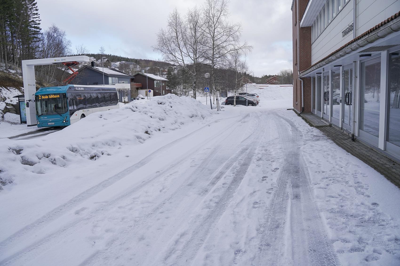 Inngangsparti ved bygg med snø, buss, vei og parkeringsplass. Foto