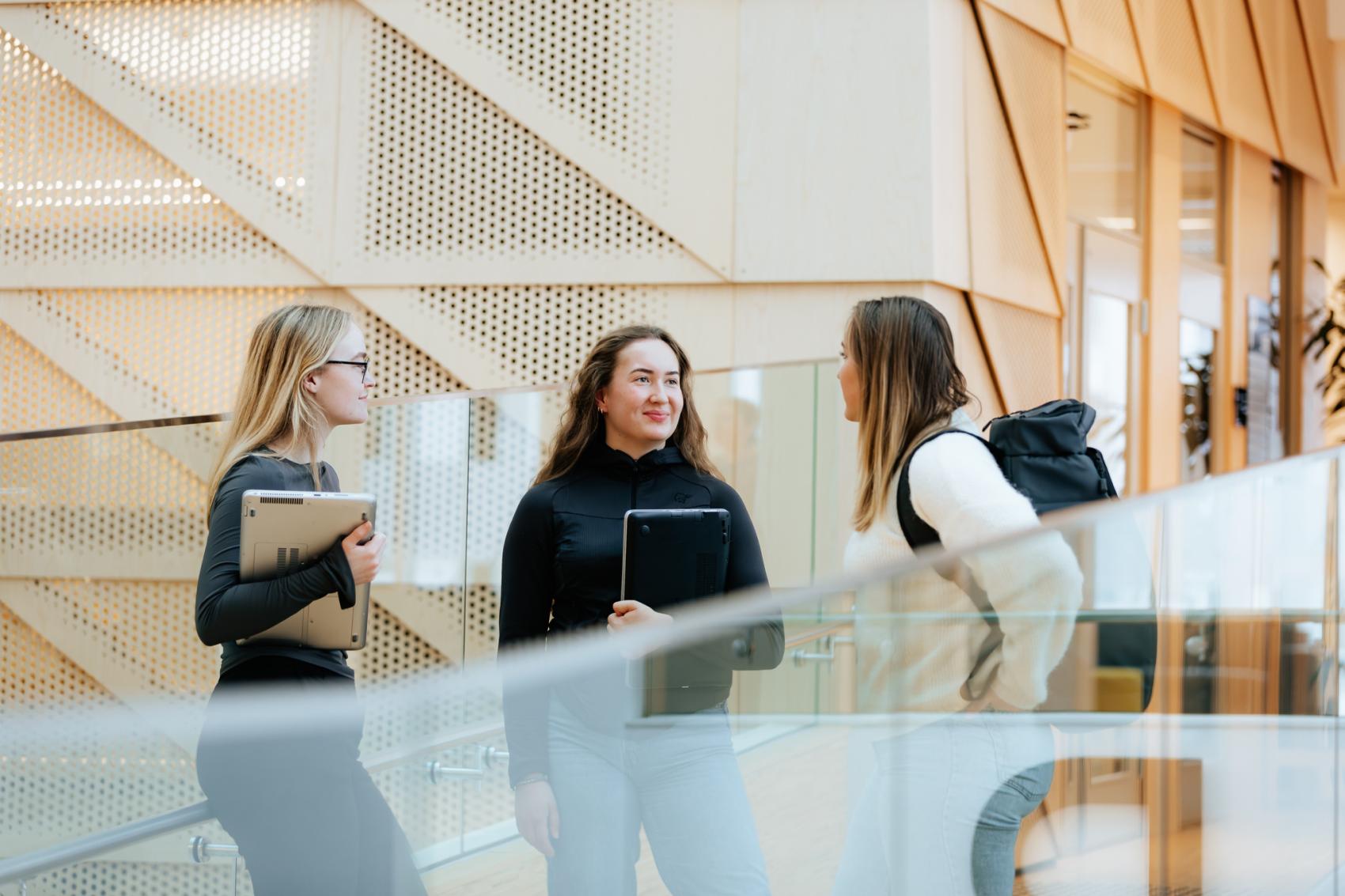 Tre kvinner som står og prater inne, med PC og sekk. Foto
