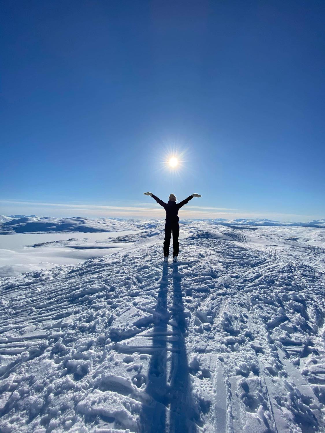 Vinterkledd person står på snødekt fjell med armene opp i lufta, vendt mot sola. Blå himmel og utsikt over flere fjelltopper i bakgrunnen.
