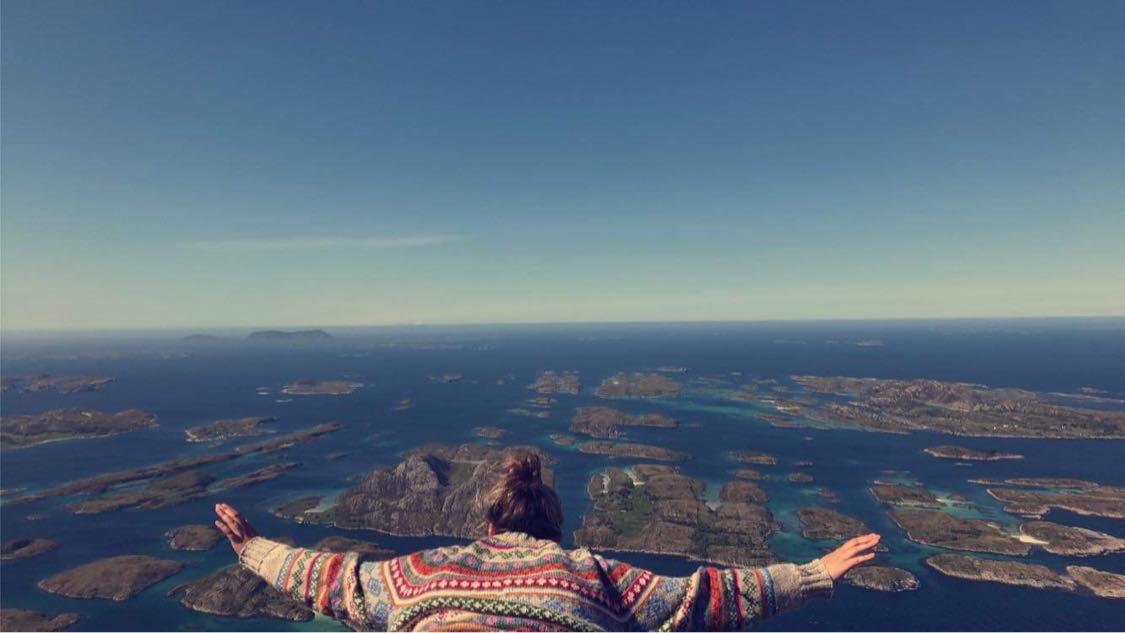 Ung dame sett bakfra, ligger på en fjelltopp med armene ut, utsikt over hav og holmer